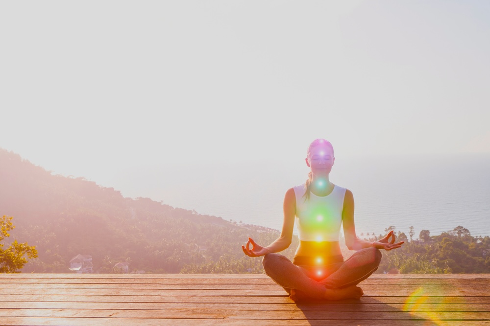 A woman meditating and sitting cross-legged on a wooden deck with glowing circles overlapped on her body to represent chakras, mountain in the background