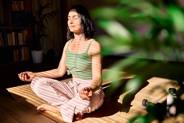 Woman practicing eight limbs of yoga and meditation at home sitting in lotus pose on yoga mat, relaxed with closed eyes.
