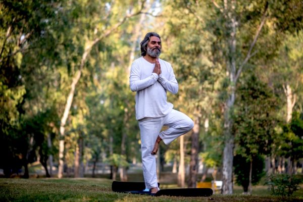 A man wearing a white pants and white shirt in a yoga pose in the park