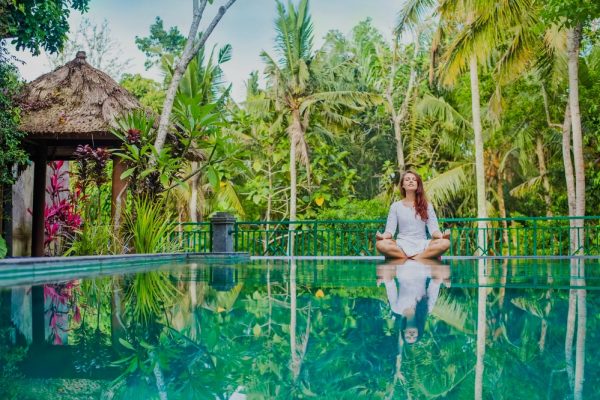 A woman meditating in front of a pool with a green hue, coconut trees, nipa hut, and nature in the background