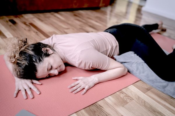 A woman is lying face down on a yoga mat, a pillow supporting one of her legs