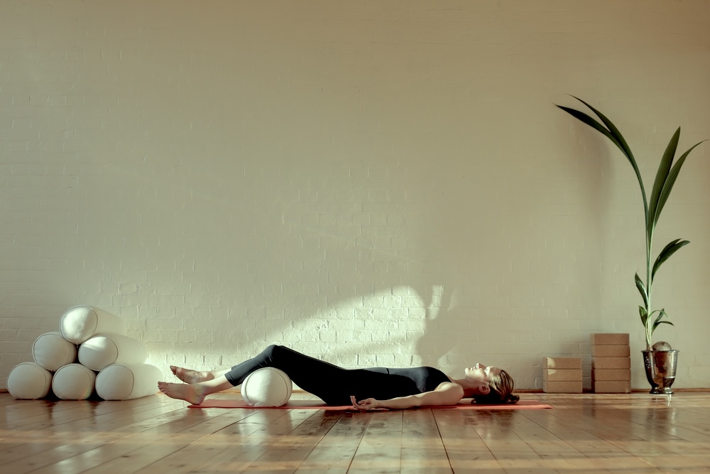 A woman lying down on a yoga mat on the floor with a pillow under her knees