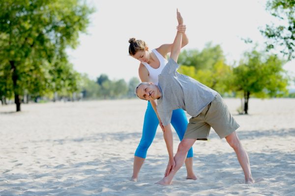 Yoga instructor assisting an elderly male on the beach