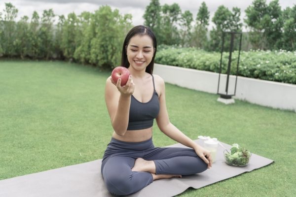 Woman sitting on a yoga mat outdoors while holding an apple