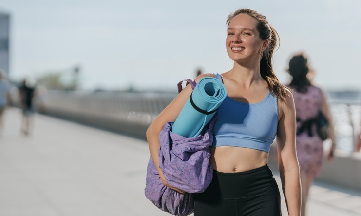 Woman walking to yoga class carrying a water bottle, yoga mat, and bag